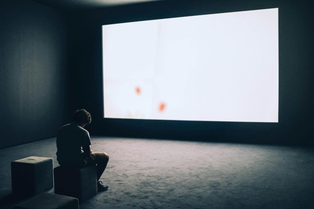 A man sits alone in a dark room with a large white screen, depicting solitude and contemplation.